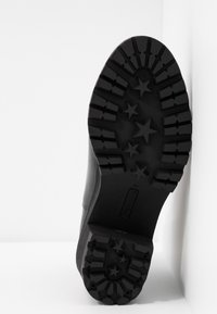 Semelle d'une botte noire avec des rainures profondes et des formes d'étoiles, inclinée contre une surface d'angle blanche.
