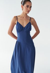 Tumši zila bezroku kleita ar V veida izgriezumu, savākta krūšu daļa un plūstoša svārku daļa. Gluda auduma ar nelielu spīdumu un minimālistisks dizains.