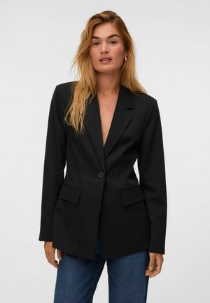 Femme aux longs cheveux blonds porte un blazer noir à un bouton et un jean bleu, debout devant un fond clair uni.