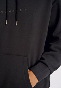 Sudadera negra de tejido suave, con bolsillo tipo canguro, capucha ajustable con cordón y logotipo en relieve en el pecho. Textura suave y puños ajustados.