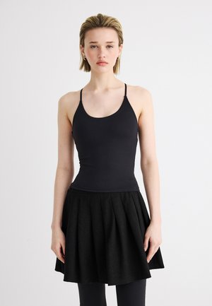 Νέα γυναίκα που φοράει μαύρο στενό αμάνικο τοπ με λεπτές τιράντες και μαύρη πλισέ φούστα μέσης, σε απλό φόντο.