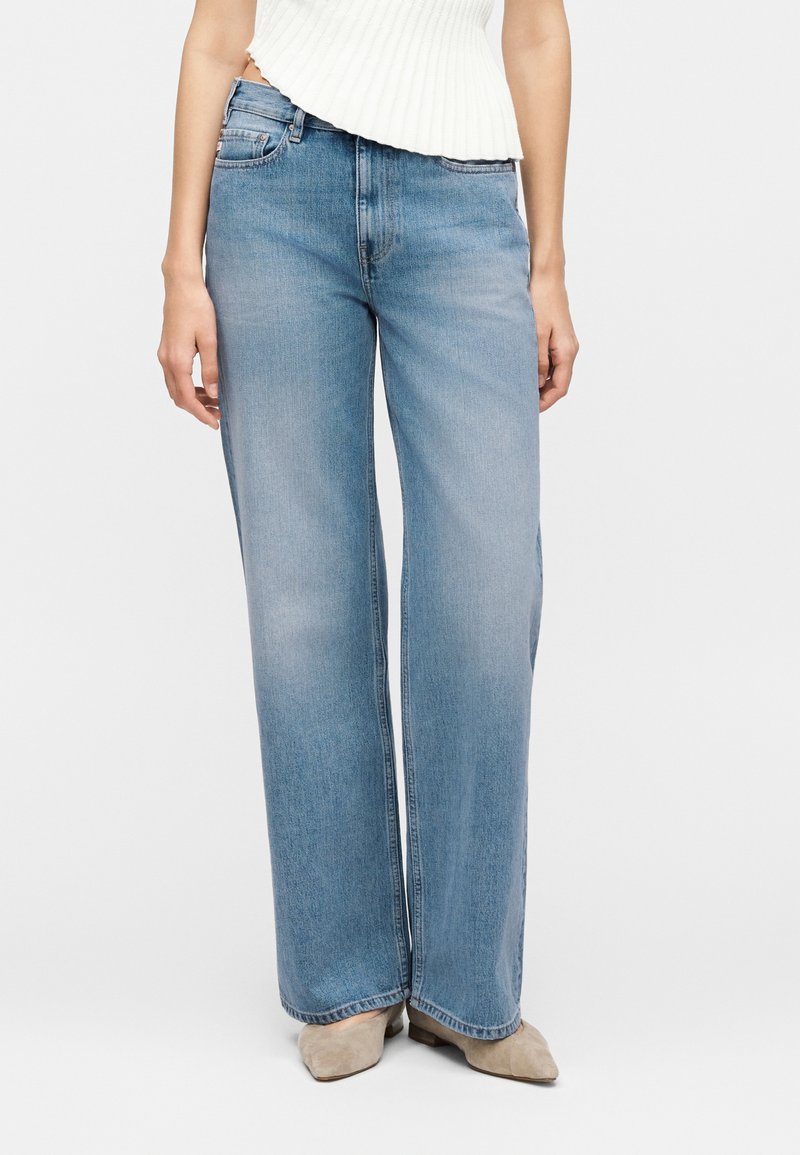 Γυναίκα που φοράει ανοιχτό μπλε τζιν ψηλόμεσο με ευρύ μπατζάκι, λευκό ριμπ αμάνικο τοπ και μπεζ μυτερά παπούτσια σε μονόχρωμο φόντο.