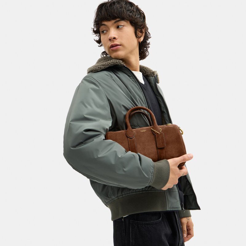 Personne en veste bomber verte tenant un petit sac à main en daim marron avec poignées, regardant sur le côté sur un fond uni.