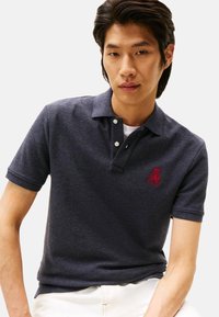 Polo de color azul marino hecho de tela texturizada, con cuello, dos botones blancos y un logotipo bordado en rojo en el lado izquierdo del pecho.