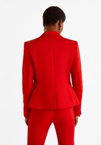 Punainen räätälöity bleiseri rakenteellisilla harteilla, peplum-yksityiskohdalla ja sileällä kankaalla. Siinä on siisti takalinja ja se on yhdistetty samansävyisiin housuihin.