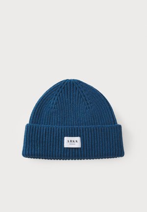 Warm blue knit beanie with ribbed texture and a folded cuff. Features a small rectangular white label with black text at the front.