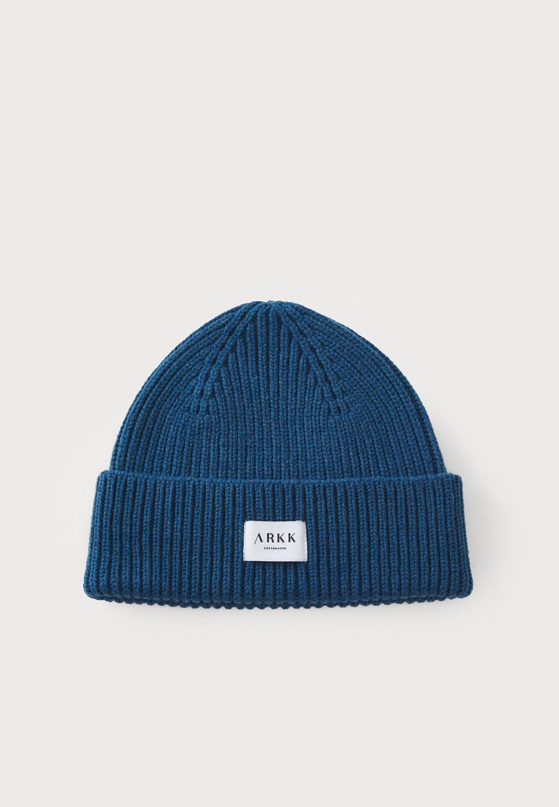 Bonnet en tricot bleu chaud avec une texture côtelée et un revers replié. Présente une petite étiquette rectangulaire blanche avec du texte noir à l'avant.
