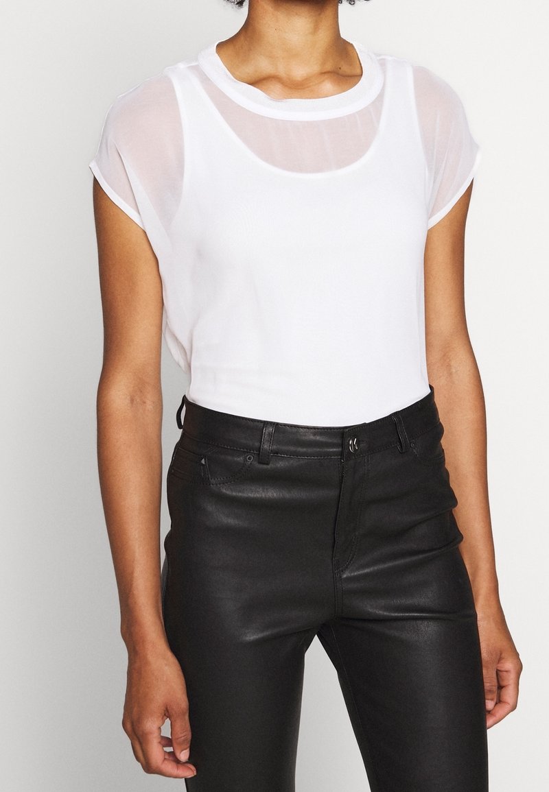 Vit kortärmad topp med en genomskinlig överdel vid halsringningen, kombinerad med svarta figurnära byxor med fickor fram och en slät yta.