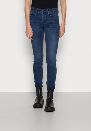 Jeans ajustados de mezclilla azul oscuro, con una cintura de altura media, cuatro bolsillos y cierre de botón. Combinados con botas negras hasta el tobillo.