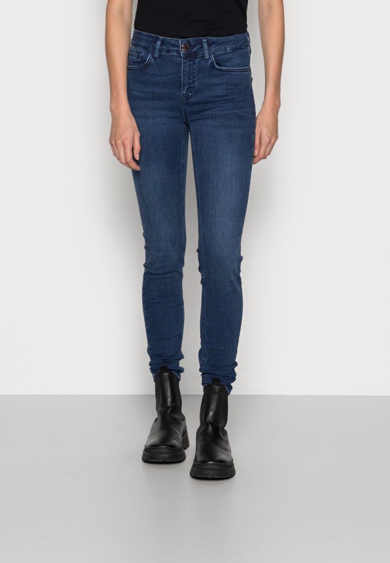 Jeans ajustados de mezclilla azul oscuro, con una cintura de altura media, cuatro bolsillos y cierre de botón. Combinados con botas negras hasta el tobillo.
