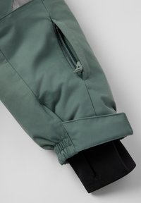 Grøn jakkeærme med lynlås-lomme, justerbar rem og sort inderskafte med tommelfingerhul på en lys baggrund.