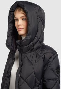 Veste doudoune noire avec une grande capuche ajustable, dotée d'un design matelassé, d'une fermeture éclair et d'un patch logo rond sur la manche.