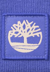 Lila tygpatch med en krämfärgad trädlogotyp omgiven av en fyrkantig kant, med en texturerad stickad bakgrund.
