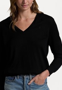 Svart V-ringad jumper i mjukt stickad tyg, med långa ärmar och en subtil broderad logotyp. Kombinerad med ljus tvättade denimjeans.