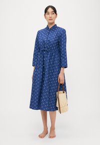 Μπλε φόρεμα με μακριά μανίκια, γιακά και ζώνη με κορδόνι στη μέση, διακοσμημένο με λευκό παϊέτ μοτίβο. Αξεσουάρ: μικρή ψάθινη τσάντα με χερούλι.