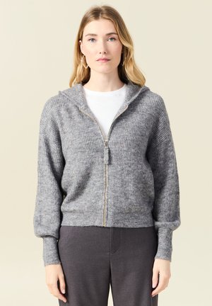 Haut à capuche zippé gris à côtes, texture douce, avec un détail de fermeture éclair doré, porté sur une chemise blanche et associé à un pantalon gris foncé.