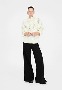 Hrubý biely pletený sveter s vysokým golierom a textúrovaným otvoreným vzorom. Doplnený o čierne široké nohavice s vysokým pásom.