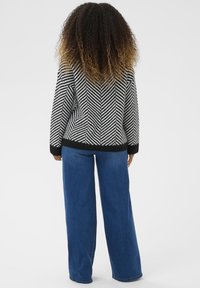 Femme avec des cheveux bouclés ombrés, portant un pull rayé noir et blanc et un pantalon bleu à jambes larges, se tenant de dos sur un fond uni.