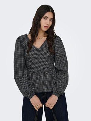 Femme aux longs cheveux foncés portant un chemisier à col en V à carreaux noirs et gris avec des manches longues bouffantes, tenant un sac noir devant elle.