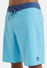 Shorts de bain bleu clair avec une taille bleu foncé et une fermeture à lacets. Texture lisse avec une étiquette logo sur la jambe inférieure gauche.