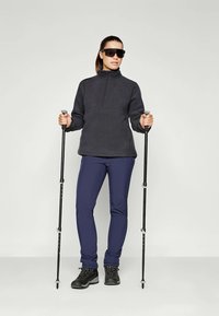 Pull en polaire gris foncé, col zippé ; associé à un pantalon de randonnée élastique bleu marine et des bottes noires imperméables. Deux bâtons de trekking à la main.