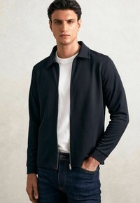 Bărbat îmbrăcat cu un sacou negru cu fermoar peste un tricou alb și blugi închiși la culoare, stând cu o mână în buzunar, pe un fundal neutr.