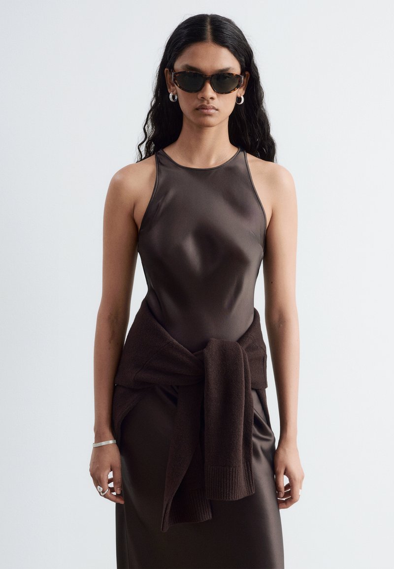 Femme aux longs cheveux noirs portant une robe en satin marron foncé sans manches, des lunettes de soleil écaille de tortue, des boucles d'oreilles créoles argentées et un pull foncé noué autour de la taille.