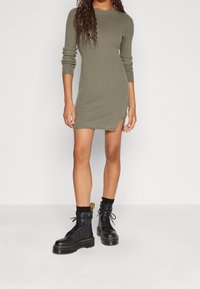 Olivgrön ribbad stickad klänning med långa ärmar och en sidoklyfta, i kombination med svarta snörstövlar med en gul logodetalj.