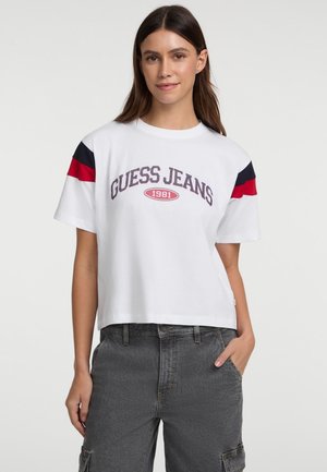 Balta trumpa marškinėlių katė su "GUESS JEANS 1981" logotipu tamsiai raudona spalva, panaudojant tamsiai mėlynas ir raudonas juosteles ant rankovių. Minkšta audinio tekstūra.
