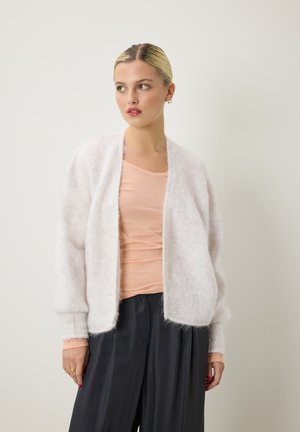Light pink, fuzzy cardigan in a soft, sheer material with ribbed cuffs. Worn over a peach top and paired with dark, loose pants.