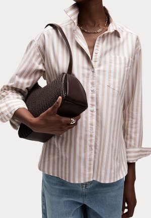 Personne portant une chemise à rayures beiges et blanches à boutons, un jean bleu, un collier en chaîne dorée, tenant un sac à main tressé marron foncé sous le bras.