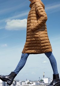 Abrigo acolchado marrón con capucha, con costuras horizontales y bolsillos. Lleva puesto sobre vaqueros azules y botines negros. Fondo urbano.