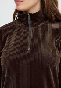 Pullover en polaire côtelé marron avec un col haut, présenté avec une courte fermeture éclair noire et une surface lisse et texturée.