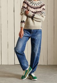 Henkilö, joka käyttää kuvioitua neuletta, sinisiä maalitahraisia farkkuja ja vihreitä tennareita seisten valkoista puuseinää vasten.