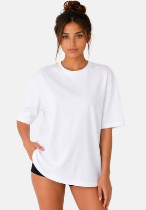 Jeune femme aux cheveux foncés attachés en chignon, portant un t-shirt blanc uni oversize et un short noir, debout devant un fond clair.