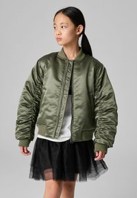 Giacca bomber verde oliva con finitura satinata, polsini a coste e tasche laterali, abbinata a una gonna in tulle nera e a un top bianco.