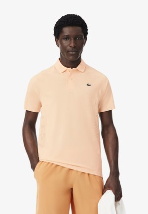 Uomo che indossa una polo arancione chiaro e pantaloncini abbinati, tiene un asciugamano bianco nella mano destra, in piedi davanti a uno sfondo neutro.