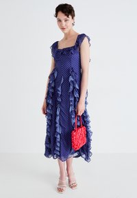Vestido de lunares morados con volantes, escote cuadrado, tejido transparente, de longitud hasta el tobillo. Bolso texturizado rojo. Modelo lleva sandalias blancas con tiras.