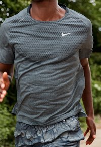 Harmaa, lyhythihainen Nike-urheilupaita, jossa on teksturoitu kuvio, pyöreä kaula-aukko ja heijastava logo, yhdistettynä kuvioituihin shortseihin.