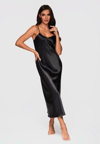 Vestido de satén negro, tipo camisa, con tirantes finos, de longitud midi, textura suave y un brillo sutil; usado con los pies descalzos.
