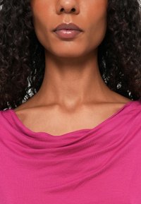 Fuksjowa bluzka z miękkim, drapowanym dekoltem, gładką fakturą i luźnym krojem. Kręcone włosy opadające na twarz na neutralnym tle.
