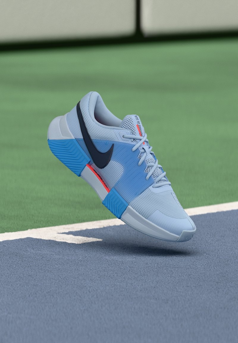 Ανοιχτό μπλε αθλητικό παπούτσι Nike με σκούρο μπλε λογότυπο swoosh, πορτοκαλί λεπτομέρειες και σχέδιο με κορδόνια, αιωρείται πάνω από την επιφάνεια ενός γηπέδου τένις.