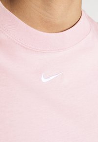 Jasnoróżowy bawełniany t-shirt z okrągłym dekoltem i małym białym logo Nike haftowanym na piersi. Gładka tekstura, prosty design.