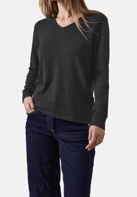 Femme portant un pull gris foncé à manches longues avec un col en V et un jean bleu foncé, debout avec une main dans la poche, sur fond blanc.