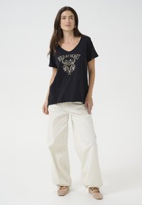 T-shirt en coton noir avec un col en V, présentant un graphique doré d'une tête de mort et de fleurs avec le texte "WILD AT HEART". Associé à un pantalon large crème.
