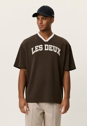 Braunes Mesh-Trikot mit kurzen Ärmeln, weißem V-Ausschnitt und dem Schriftzug "LES DEUX" auf der Vorderseite. Kombiniert mit hellfarbigen Hosen.