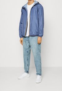 Blå mønsterjakke med elastiske manchetter og hætte, åbnet over en hvid t-shirt, parret med lyseblå denimjeans og hvide sko.