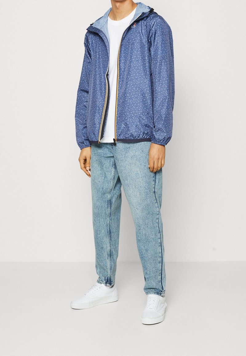 Blå mønsterjakke med elastiske manchetter og hætte, åbnet over en hvid t-shirt, parret med lyseblå denimjeans og hvide sko.