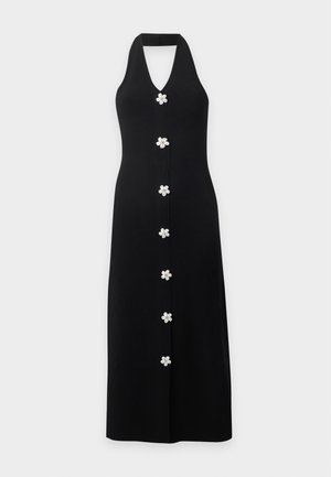 Femme portant une robe noire sans manches à col halter avec des boutons blancs en forme de fleurs sur le devant, associée à des talons hauts de couleur claire.