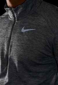 Pullover half-zip Nike in tessuto grigio scuro testurizzato con logo riflettente argento sul petto, dotato di colletto alto e chiusura con zip.